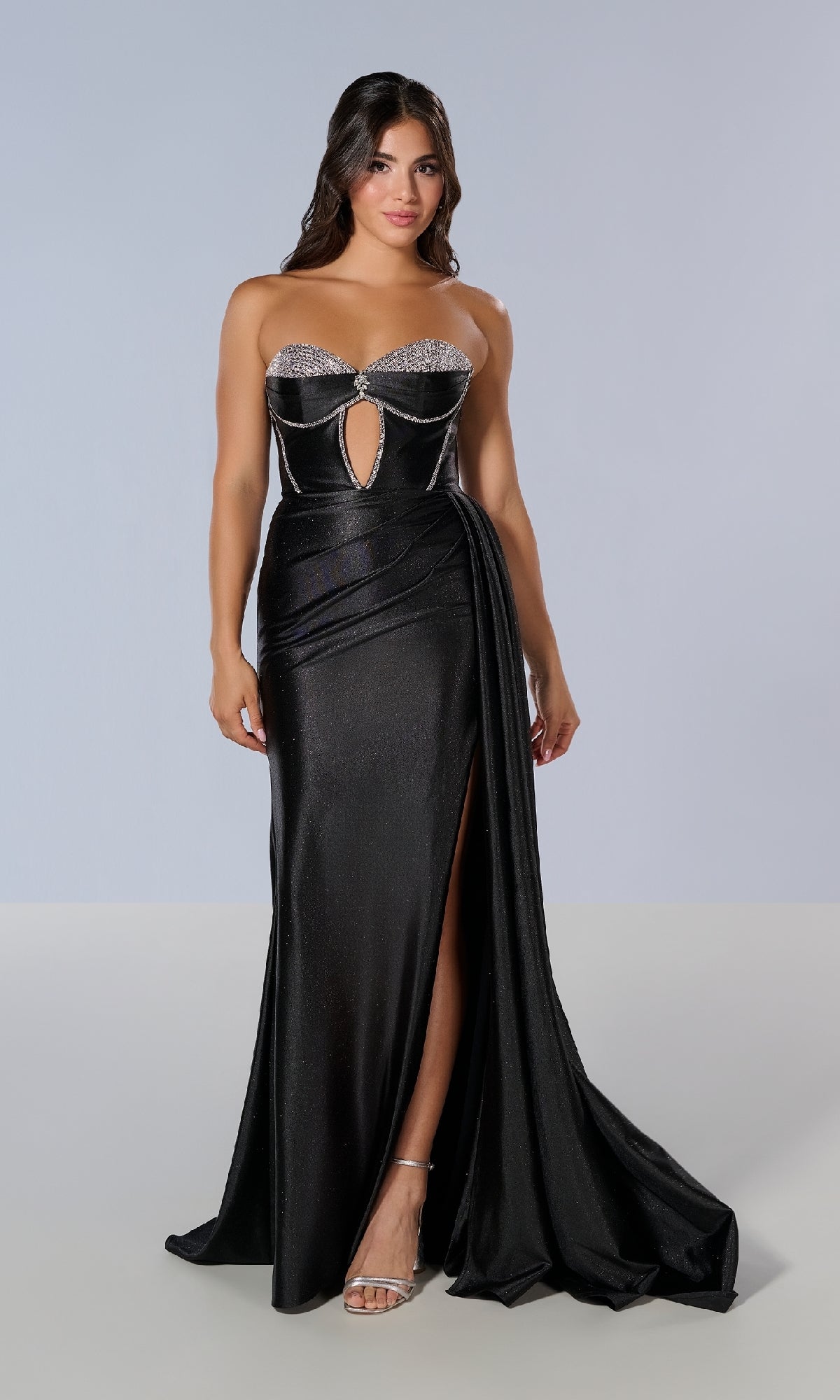 A woman wears the Tiffany 16197 Long Prom Dress—a strapless black gown with a silver embellished bust, cut-out corset bodice, high slit, and draped side detail—standing against a plain background.
